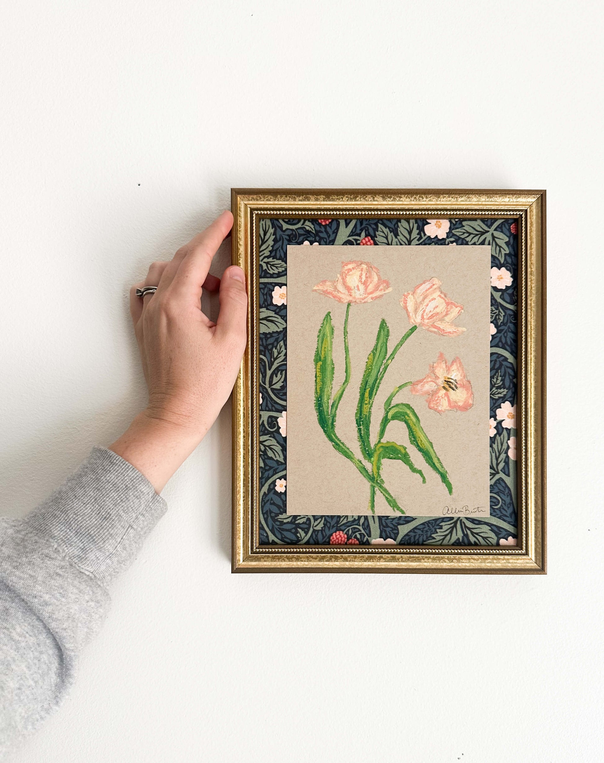 peach florals painted with oil pastels on tan paper, mounted on floral fabric and framed in a gold frame. Painting shown on a white wall