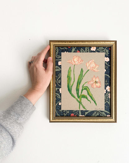 peach florals painted with oil pastels on tan paper, mounted on floral fabric and framed in a gold frame. Painting shown on a white wall