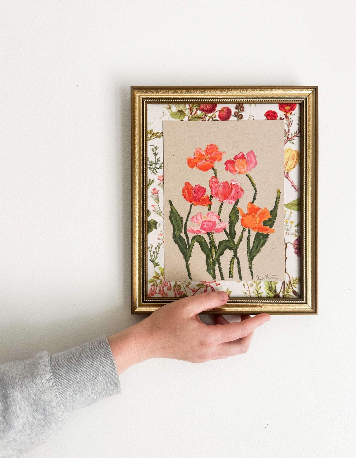 A painting of pink and orange flowers on tan paper mounted on floral fabric. The painting is in a gold frame and is shown on a white wall.