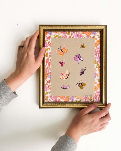 Eight colorful butterflies painted on tan toned paper and mounted on colorful, floral fabric. The painting is framed in a gold frame and shown on a white wall.