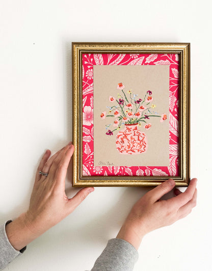 A pink vase of small flowers painted on tan paper and mounted on pink floral fabric. The painting is framed in a gold frame and shown on a white wall.