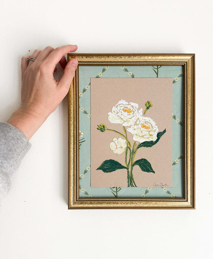 A painting of white peonies on tan paper mounted on floral fabric. The painting is in a gold frame and shown on a white wall.