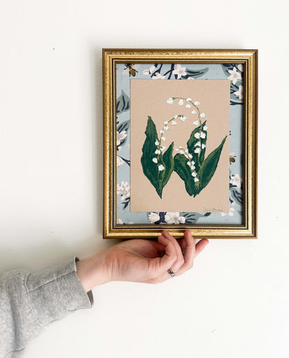 painting of lily of the valley flower on tan toned paper, framed in a gold frame and mounted on a blue floral fabric