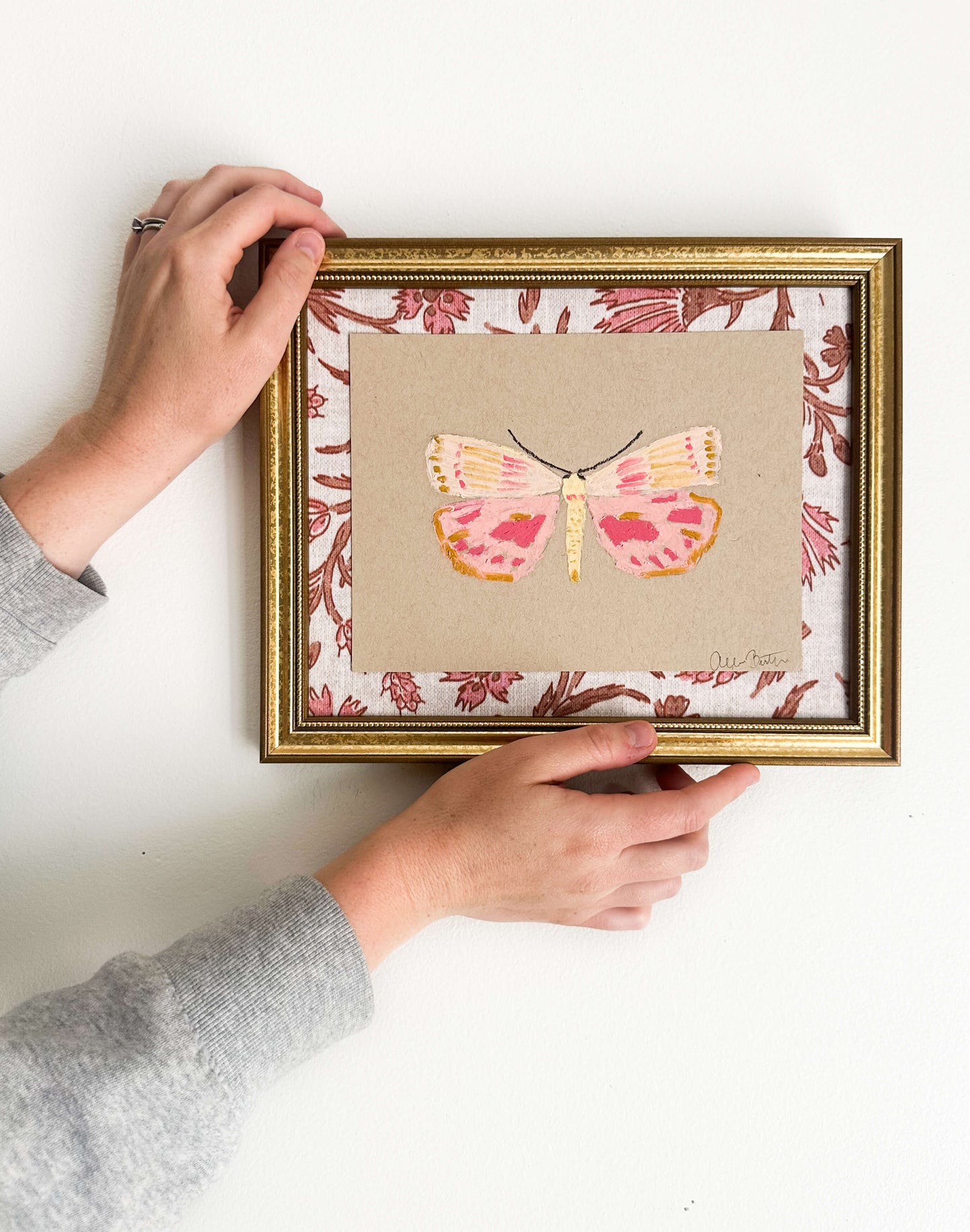 a pink and yellow butterfly painting on tan paper, mounted on pink and tan floral fabric and framed in a gold frame. Painting is shown on a white wall.