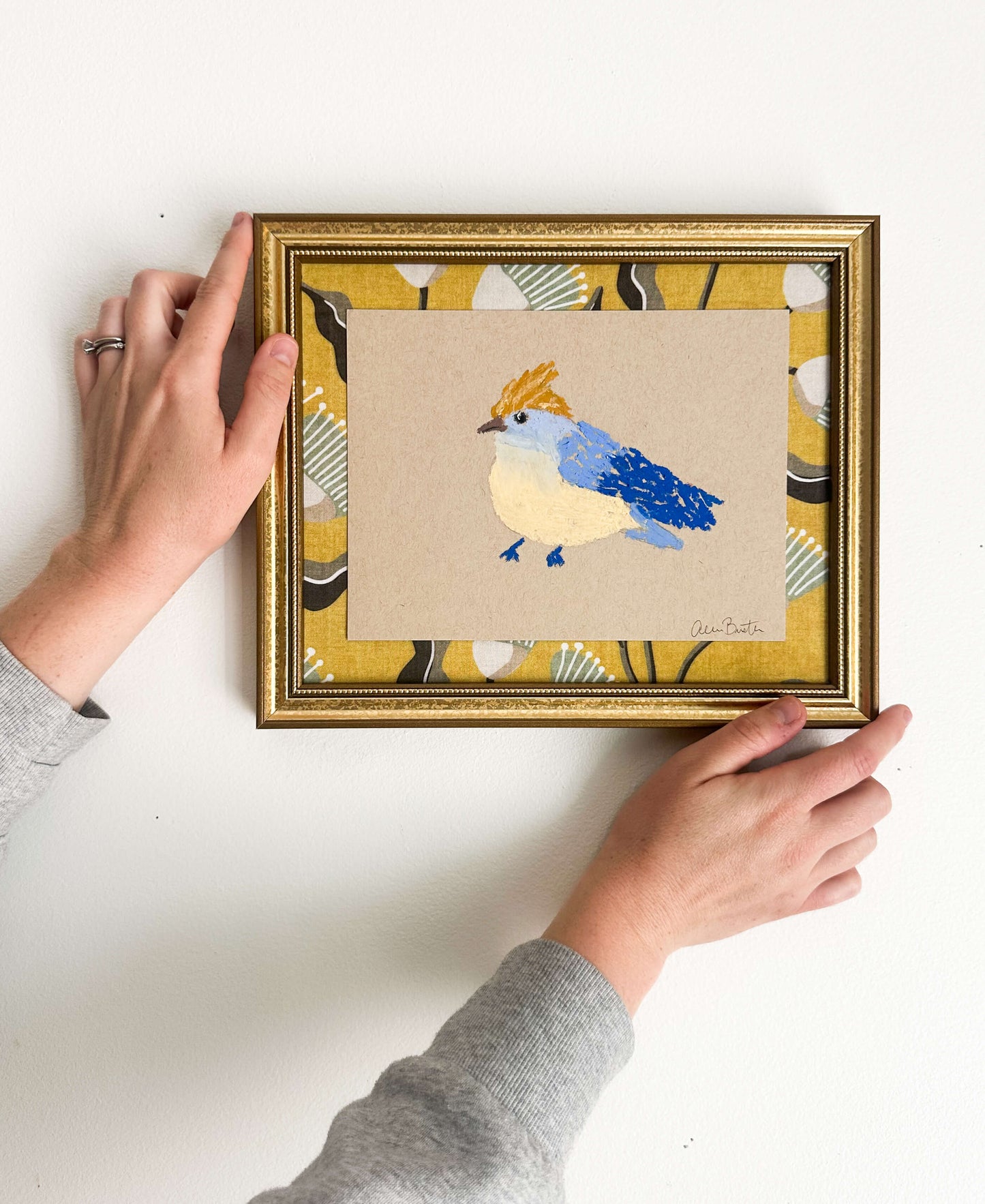 a bird painting with blue and yellow oil pastels on tan paper, mounted on yellow fabric and framed in a gold frame shown on a white wall