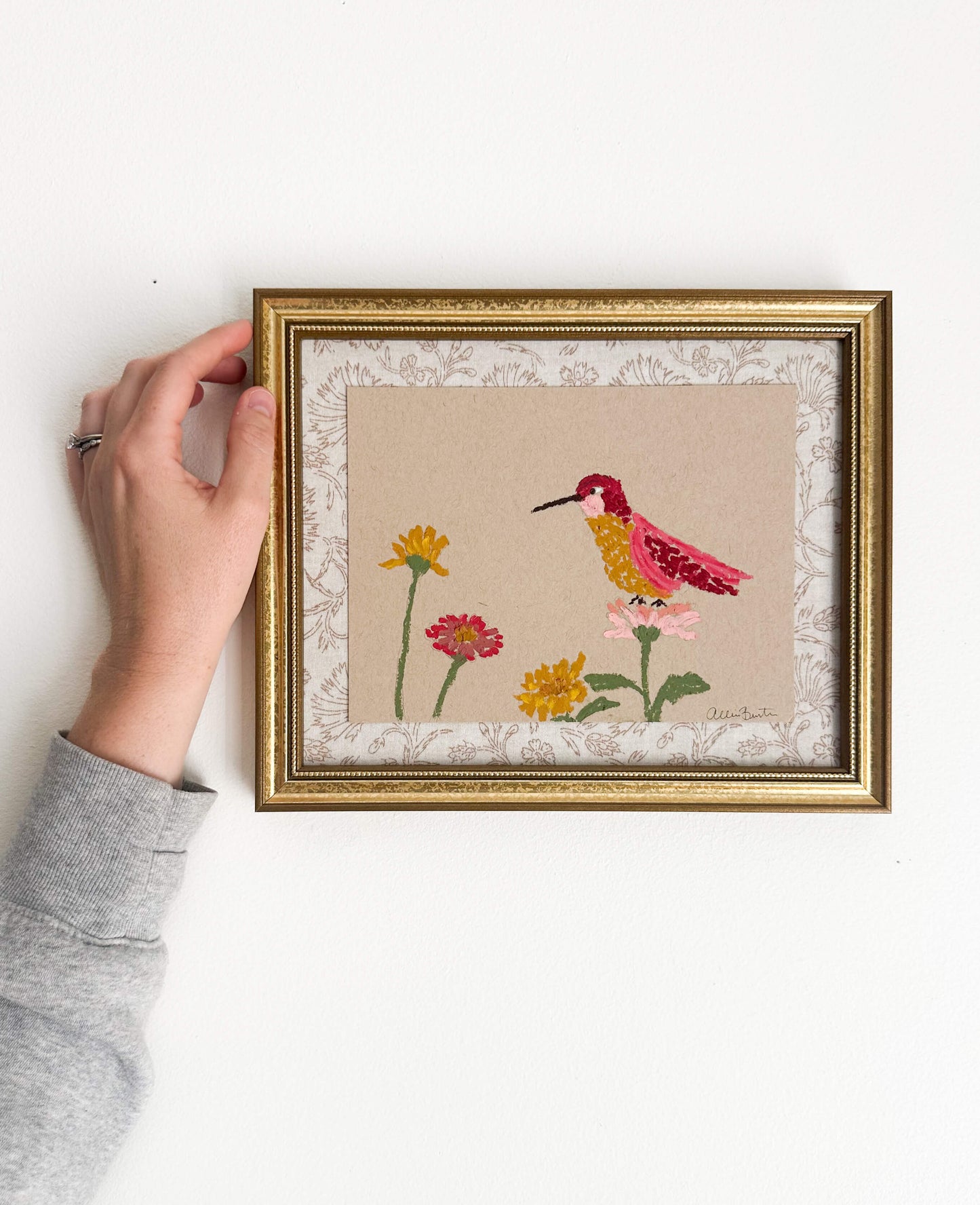 A painting of a colorful hummingbird and flowers on tan paper mounted on tan floral fabric. The painting is in a gold frame and is shown on a white wall.