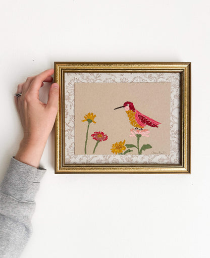 A painting of a colorful hummingbird and flowers on tan paper mounted on tan floral fabric. The painting is in a gold frame and is shown on a white wall.