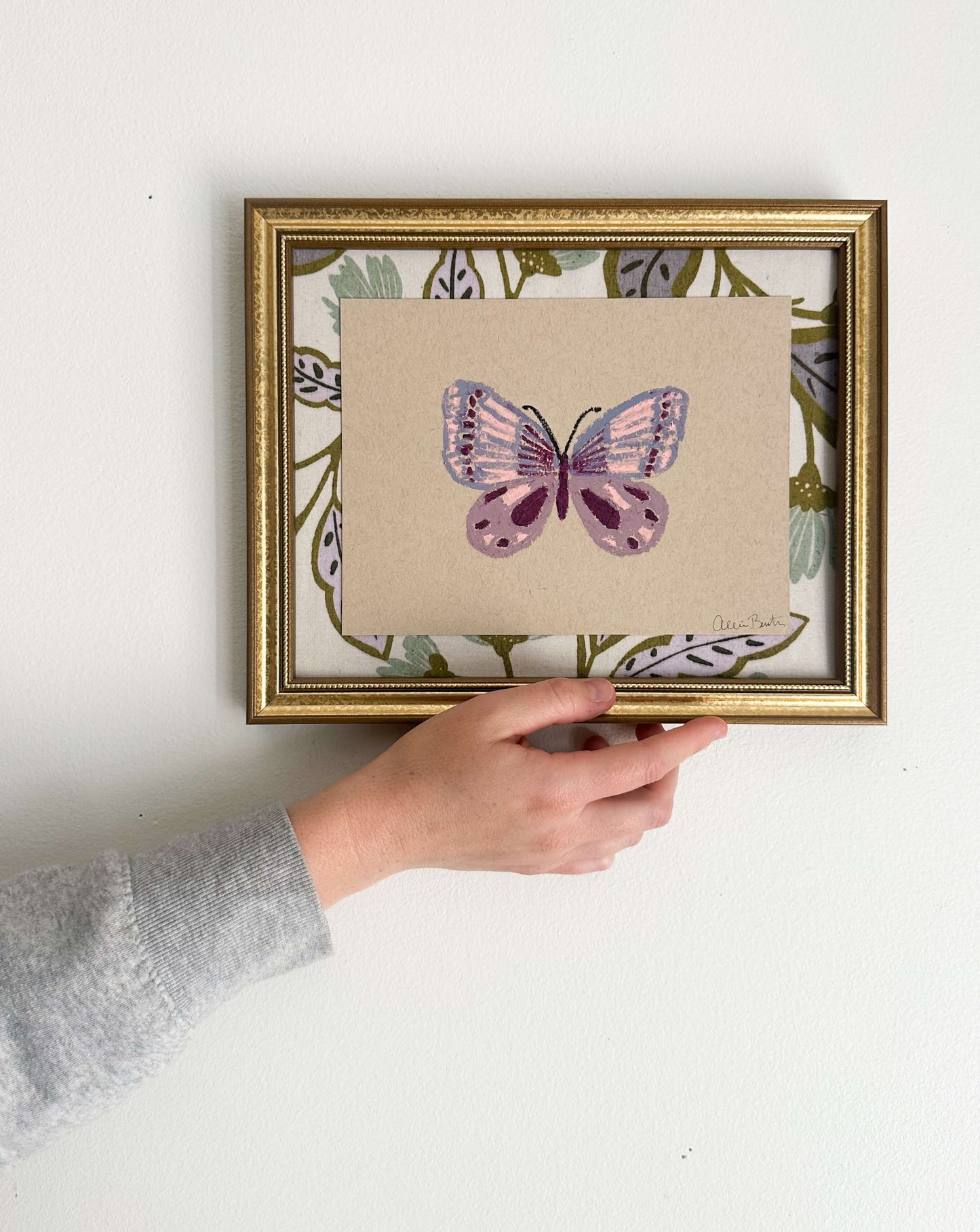 A painting of a purple butterfly on tan paper and mounted on floral fabric. The painting is in a gold frame and shown on a white wall.