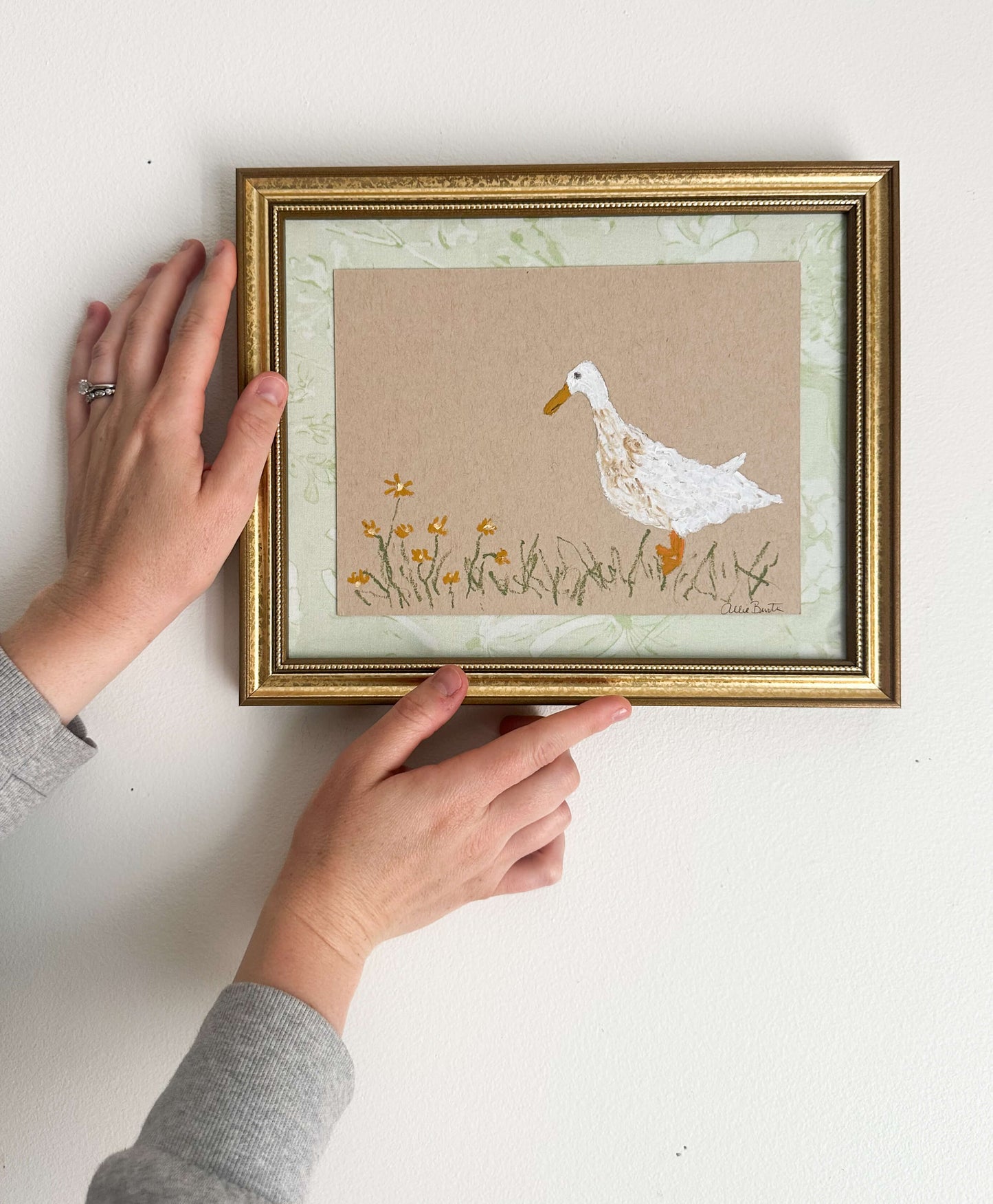 A painting of a goose with flowers on tan paper mounted on light green fabric. The painting is in a gold frame on a white wall.