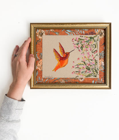 A painting of an orange hummingbird and pink flowers on tan paper. The painting is in a gold frame on a white wall.