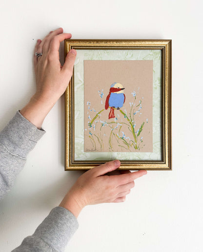 A painting of a colorful bird perched on a plant painted on tan paper and mounted on light green fabric. The painting is in a gold frame and shown on a white wall