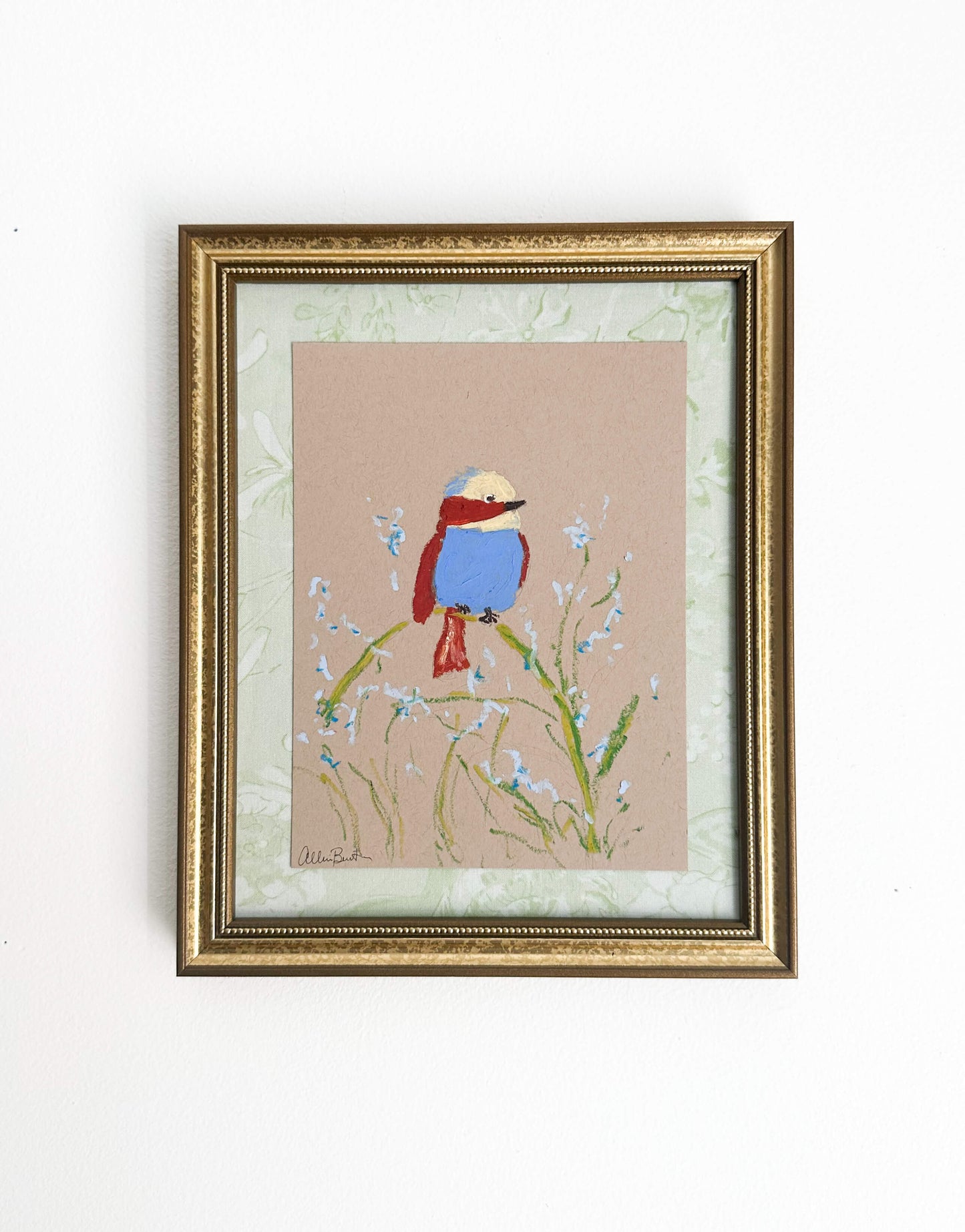 A painting of a colorful bird perched on a plant painted on tan paper and mounted on light green fabric. The painting is in a gold frame and shown on a white wall