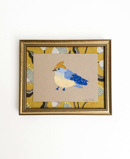 a bird painting with blue and yellow oil pastels on tan paper, mounted on yellow fabric and framed in a gold frame shown on a white wall