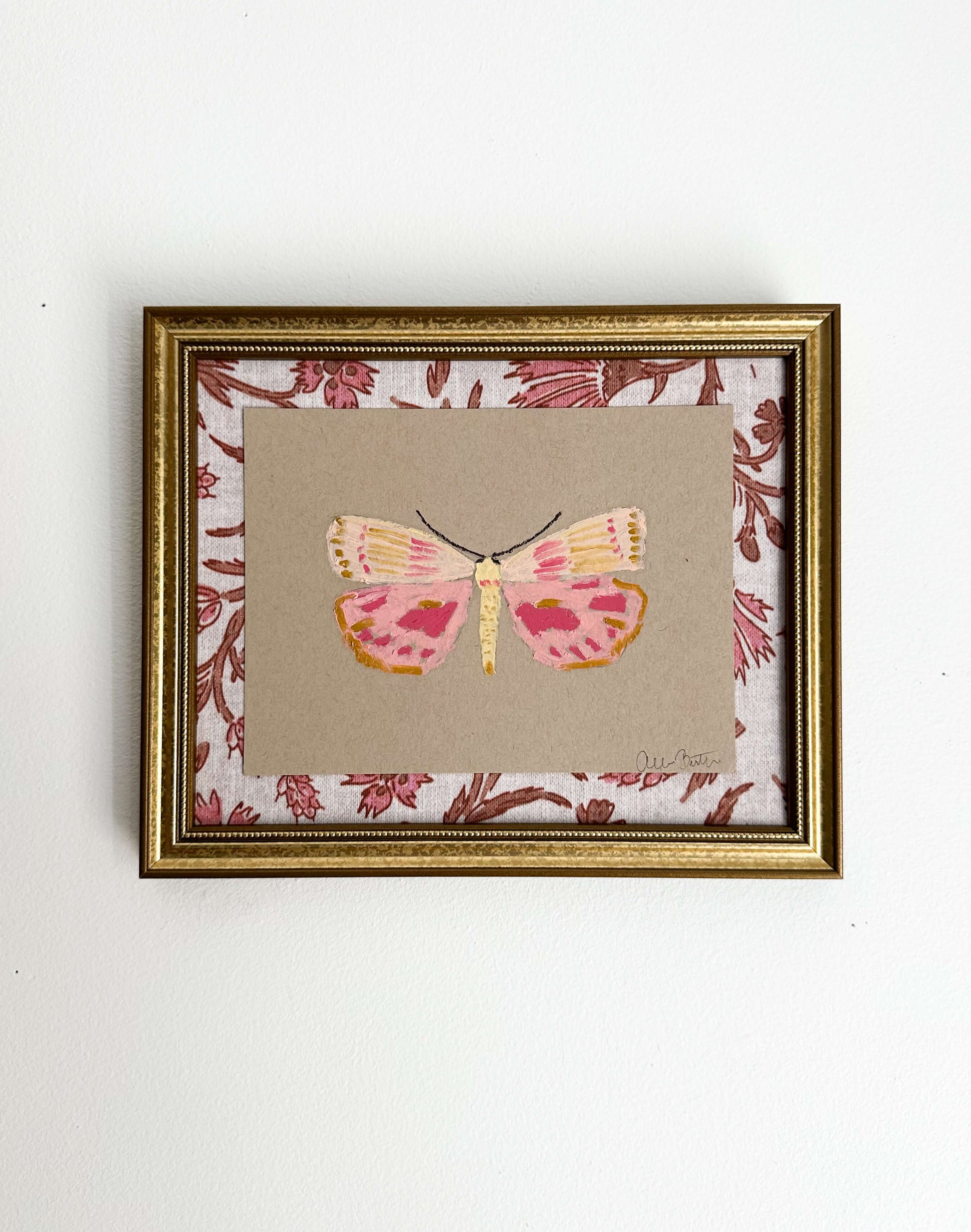 a pink and yellow butterfly painting on tan paper, mounted on pink and tan floral fabric and framed in a gold frame. Painting is shown on a white wall.