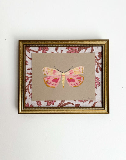 a pink and yellow butterfly painting on tan paper, mounted on pink and tan floral fabric and framed in a gold frame. Painting is shown on a white wall.