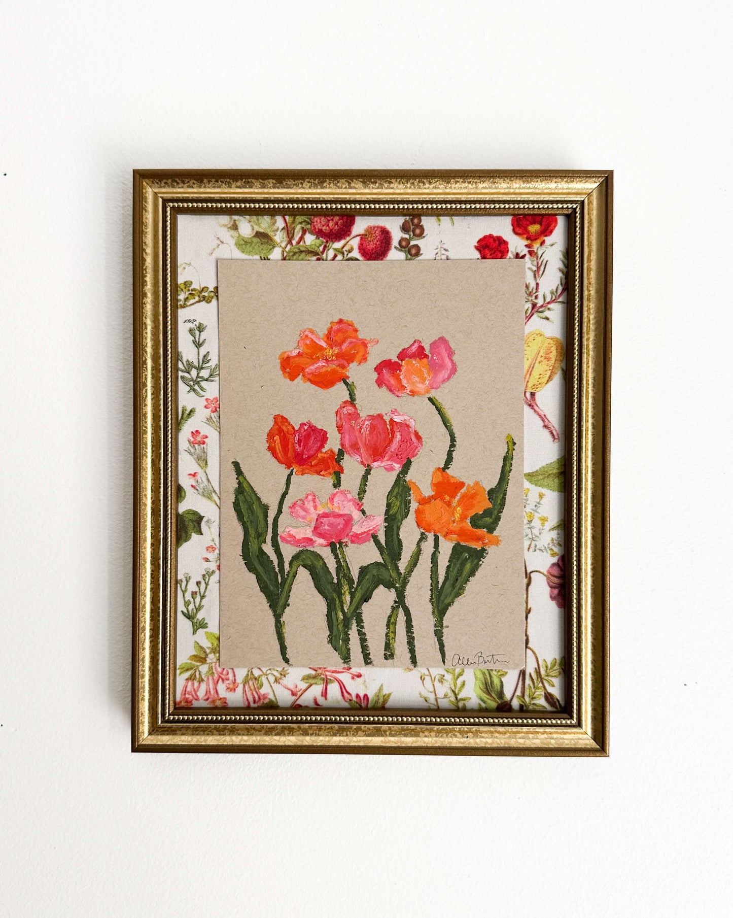 A painting of pink and orange flowers on tan paper mounted on floral fabric. The painting is in a gold frame and is shown on a white wall.