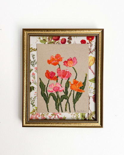 A painting of pink and orange flowers on tan paper mounted on floral fabric. The painting is in a gold frame and is shown on a white wall.