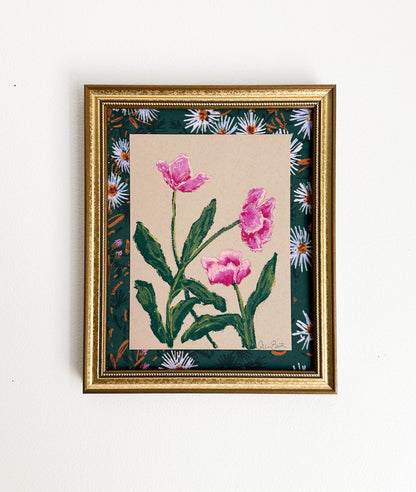 pink flowers painted on tan paper and mounted on floral fabric in a gold frame. The painting is shown on a white wall.