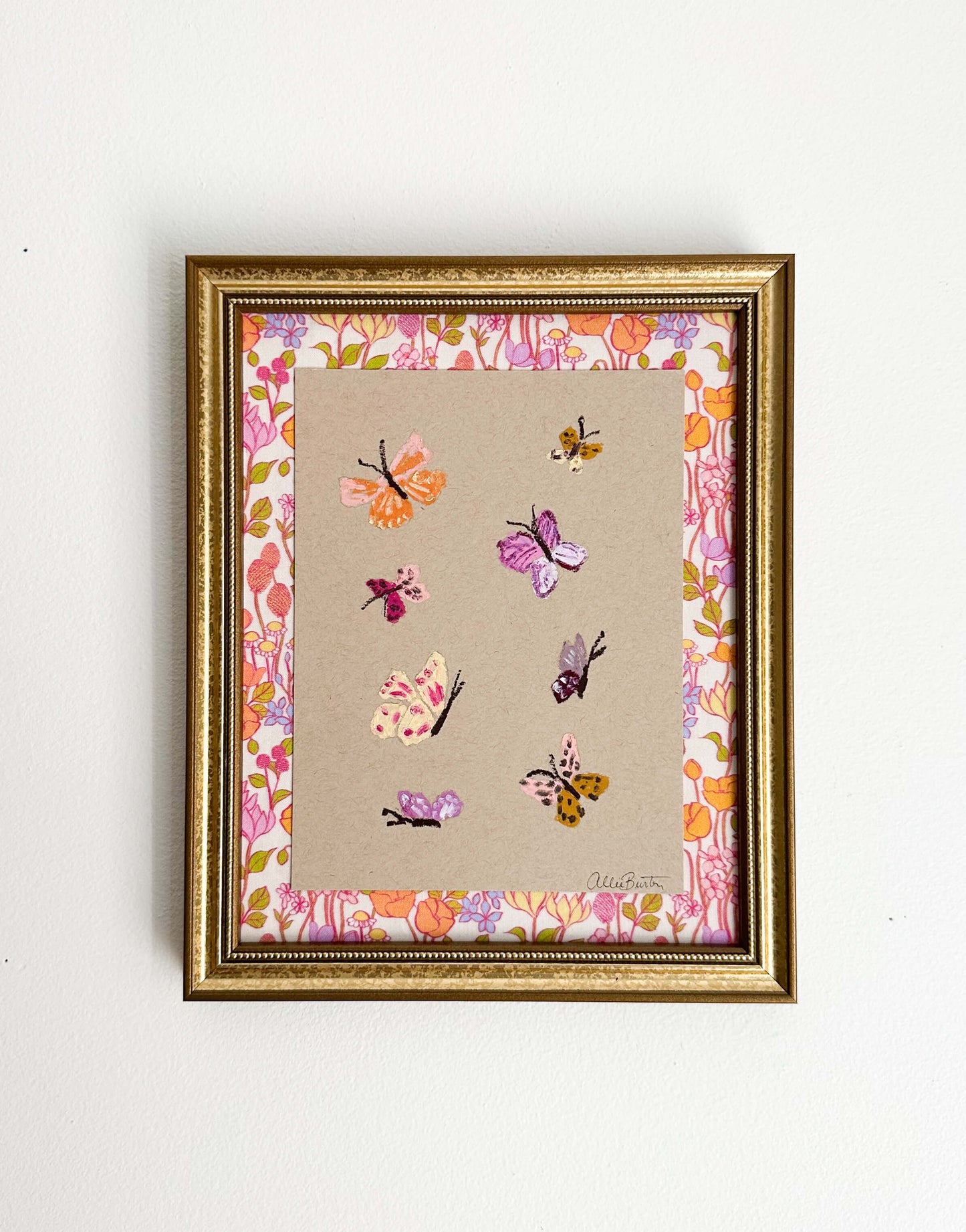 Eight colorful butterflies painted on tan toned paper and mounted on colorful, floral fabric. The painting is framed in a gold frame and shown on a white wall.