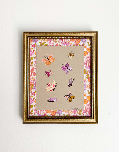 Eight colorful butterflies painted on tan toned paper and mounted on colorful, floral fabric. The painting is framed in a gold frame and shown on a white wall.