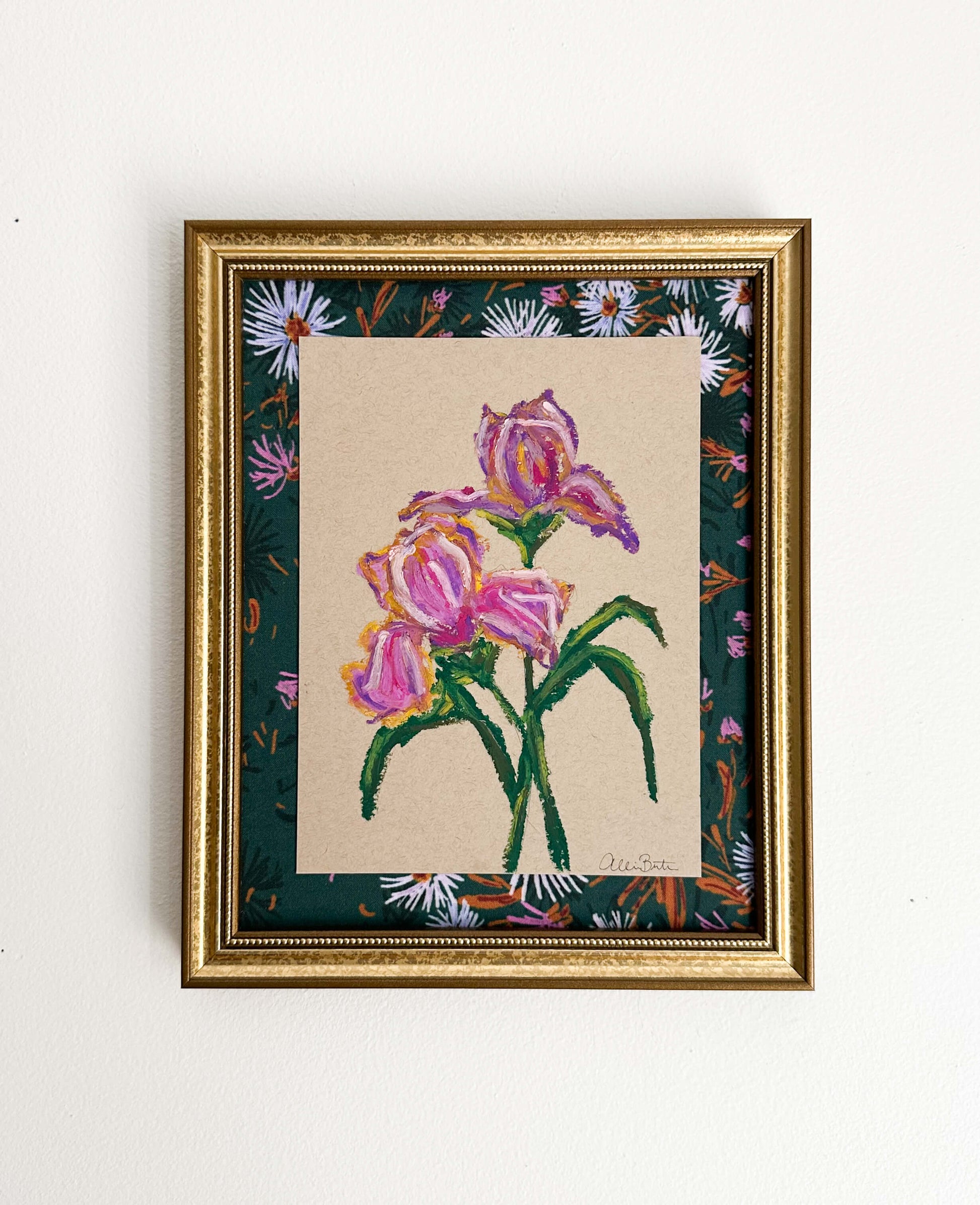A painting of purple irises on tan paper mounted on dark floral fabric. The painting is in a gold frame and shown on a white wall.