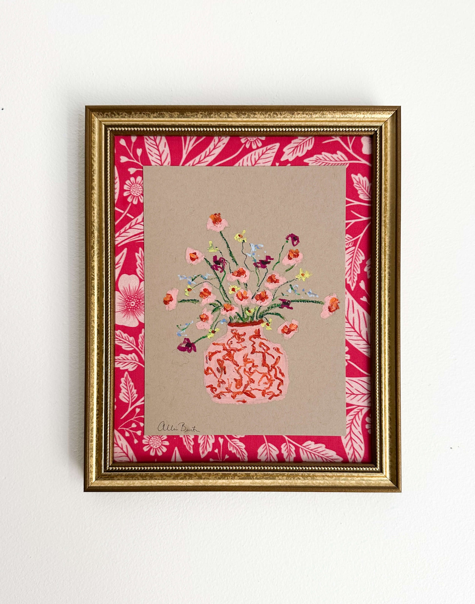 A pink vase of small flowers painted on tan paper and mounted on pink floral fabric. The painting is framed in a gold frame and shown on a white wall.