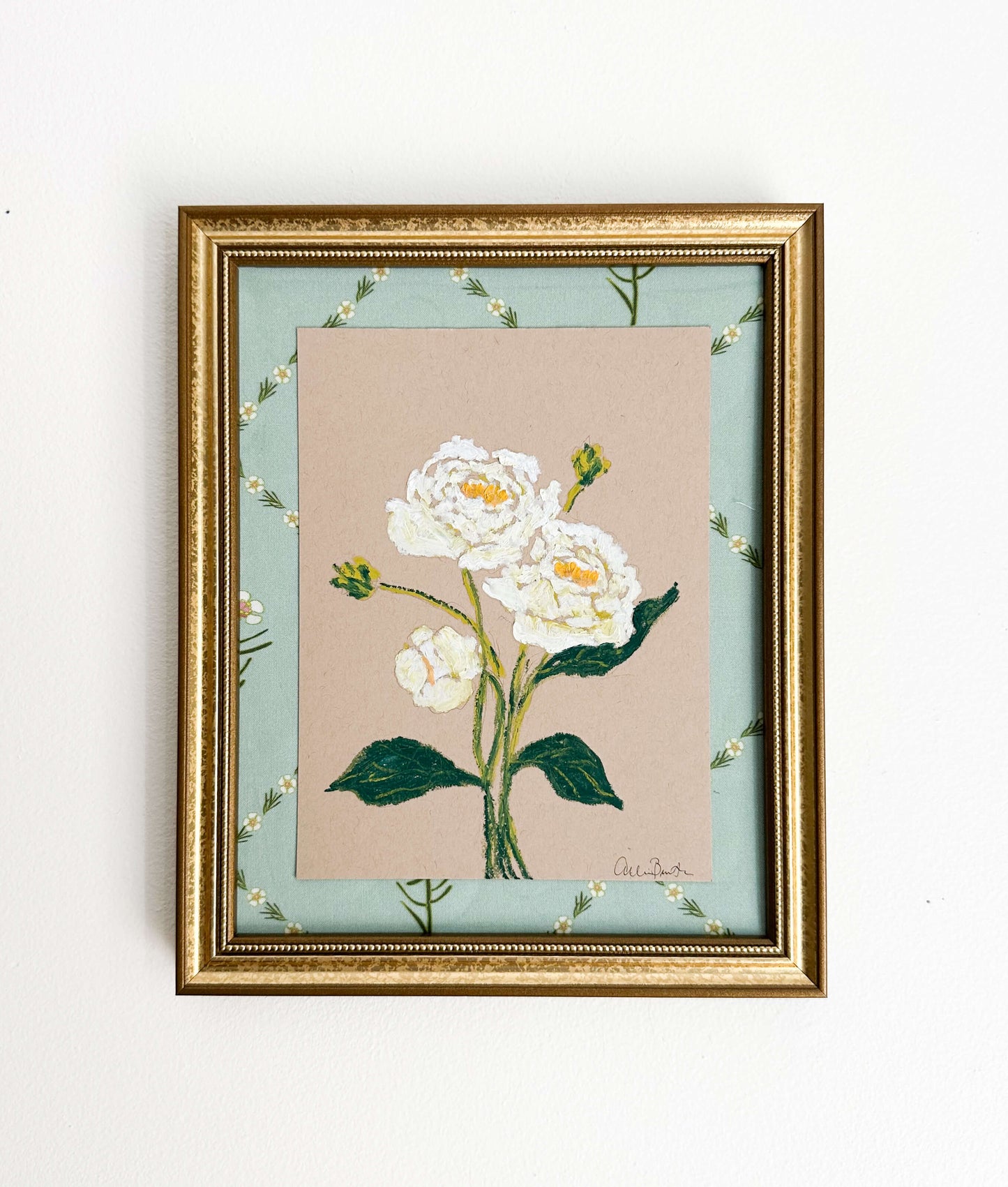 A painting of white peonies on tan paper mounted on floral fabric. The painting is in a gold frame and shown on a white wall.