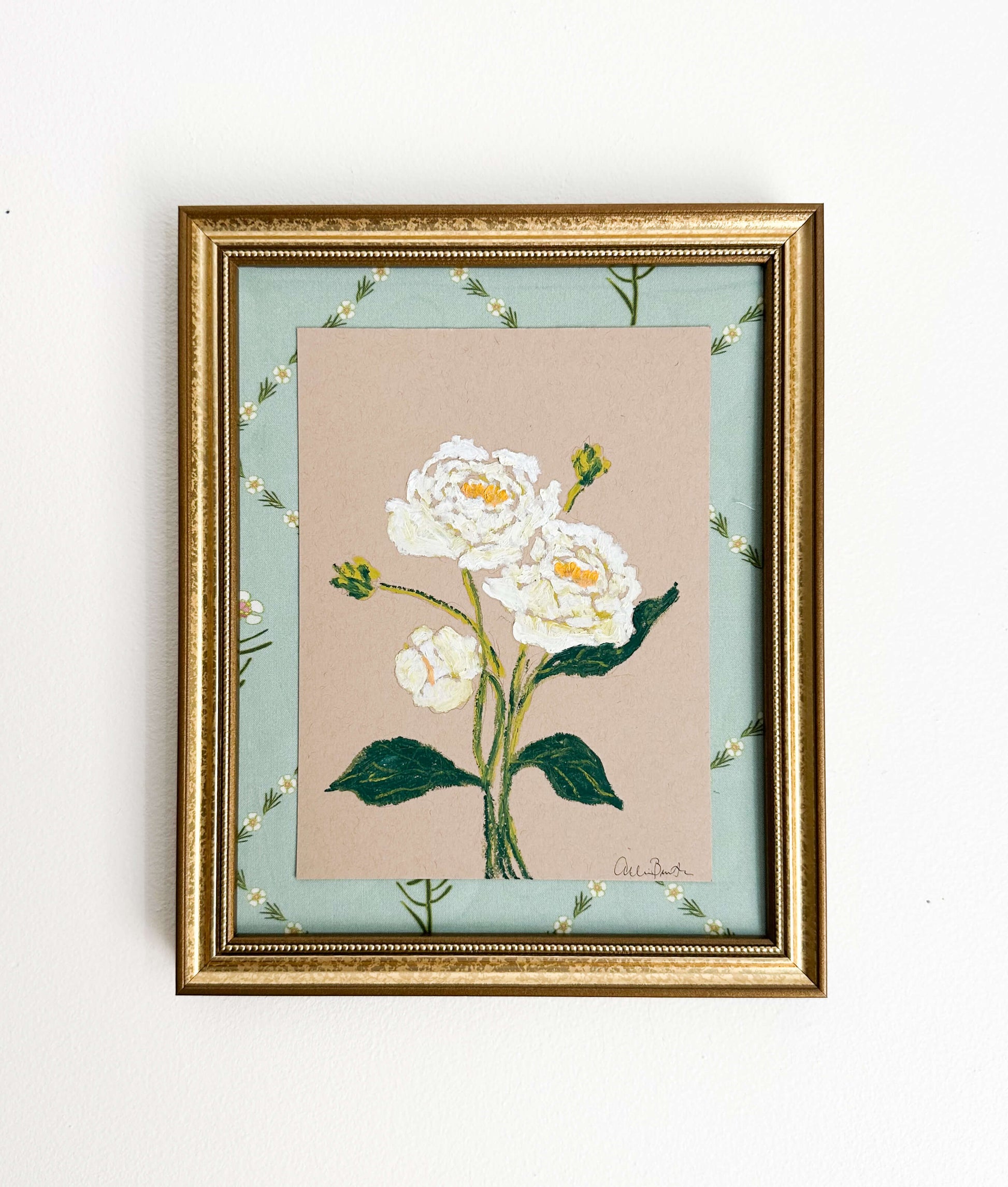 A painting of white peonies on tan paper mounted on floral fabric. The painting is in a gold frame and shown on a white wall.