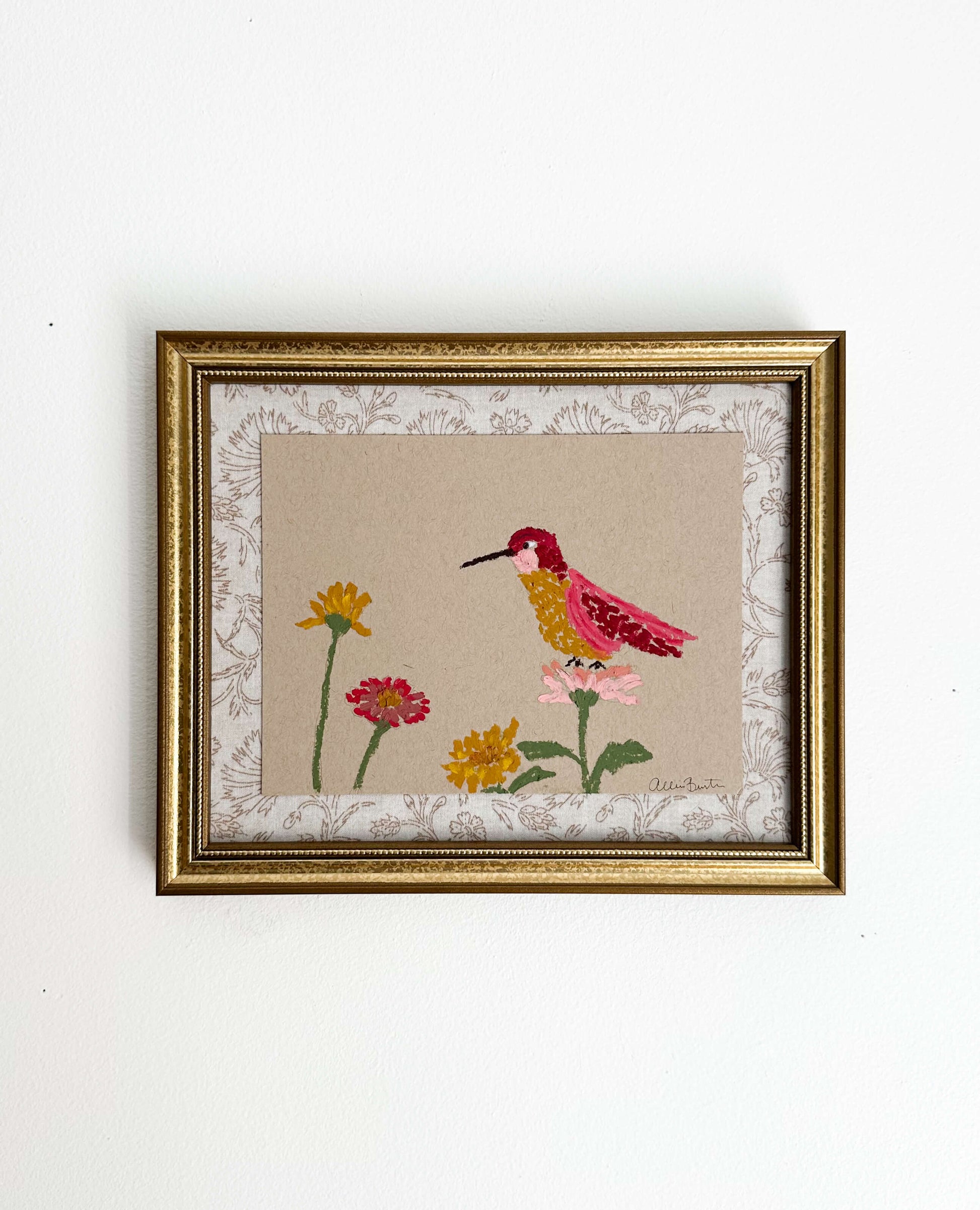 A painting of a colorful hummingbird and flowers on tan paper mounted on tan floral fabric. The painting is in a gold frame and is shown on a white wall.