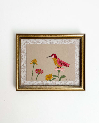 A painting of a colorful hummingbird and flowers on tan paper mounted on tan floral fabric. The painting is in a gold frame and is shown on a white wall.