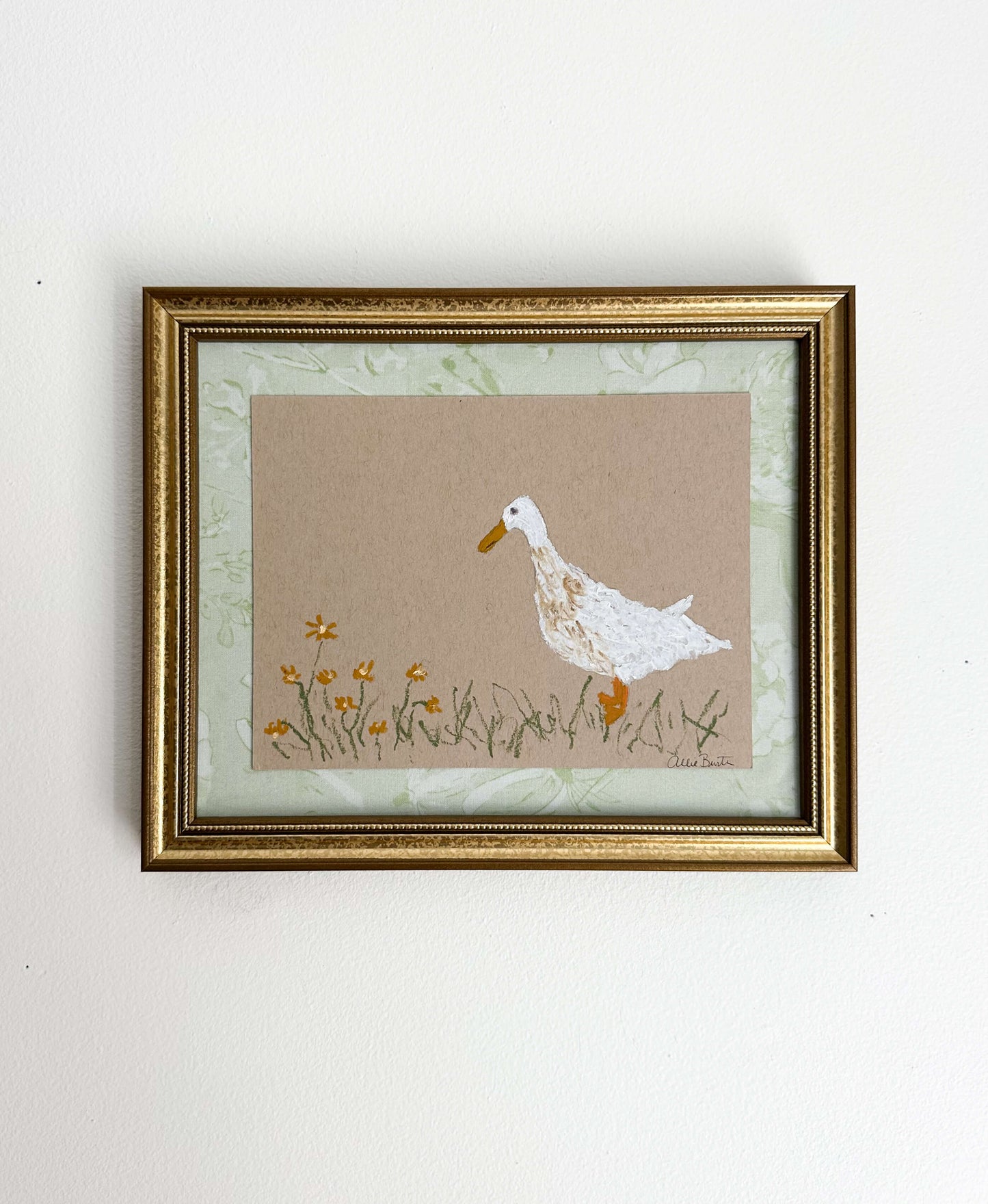 A painting of a goose with flowers on tan paper mounted on light green fabric. The painting is in a gold frame on a white wall.