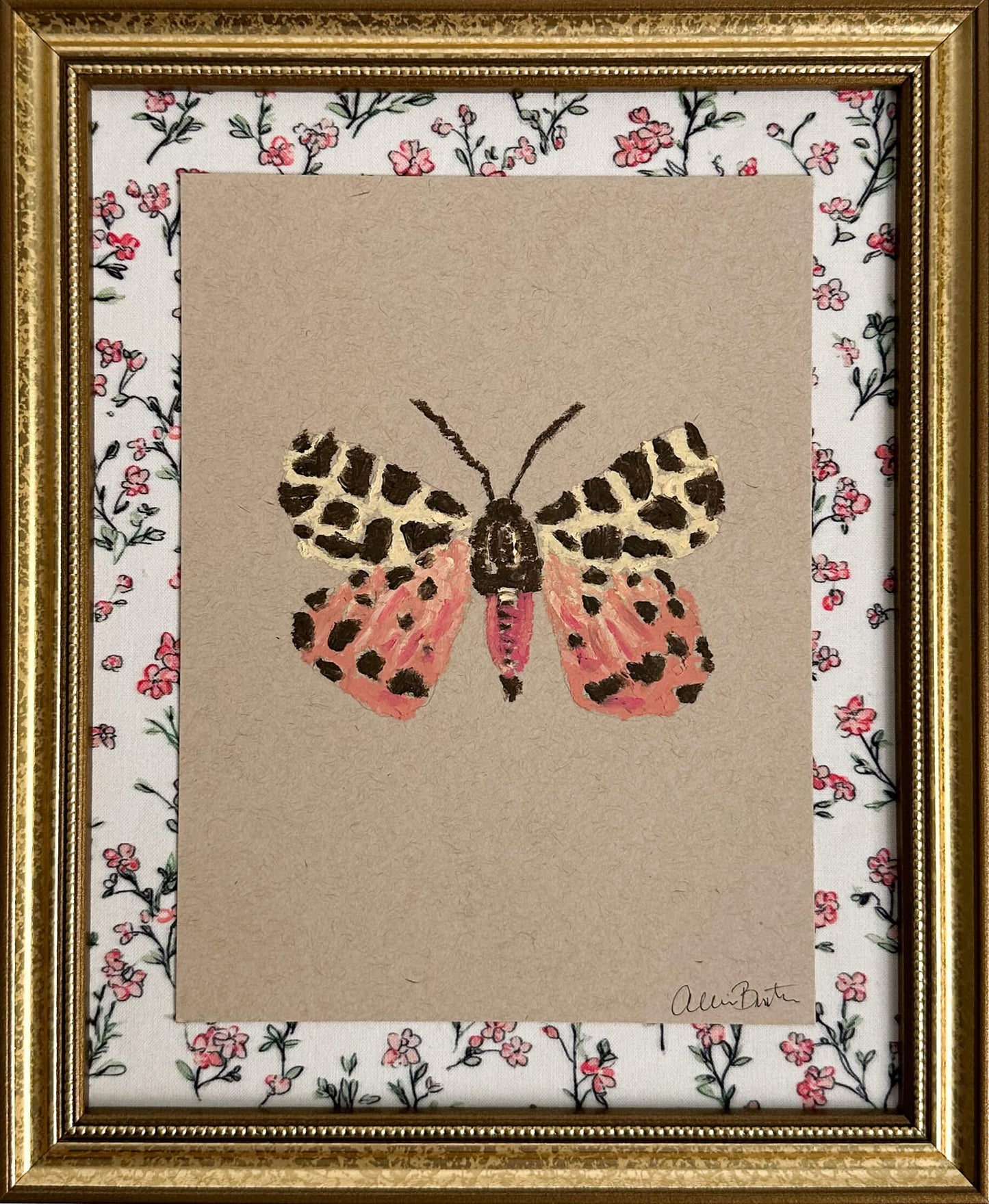 a pink, yellow and brown moth painted on tan paper. The paper is mounted on floral fabric and framed in a gold frame