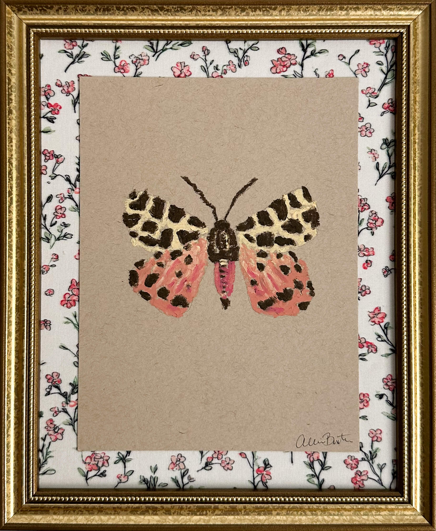 a pink, yellow and brown moth painted on tan paper. The paper is mounted on floral fabric and framed in a gold frame