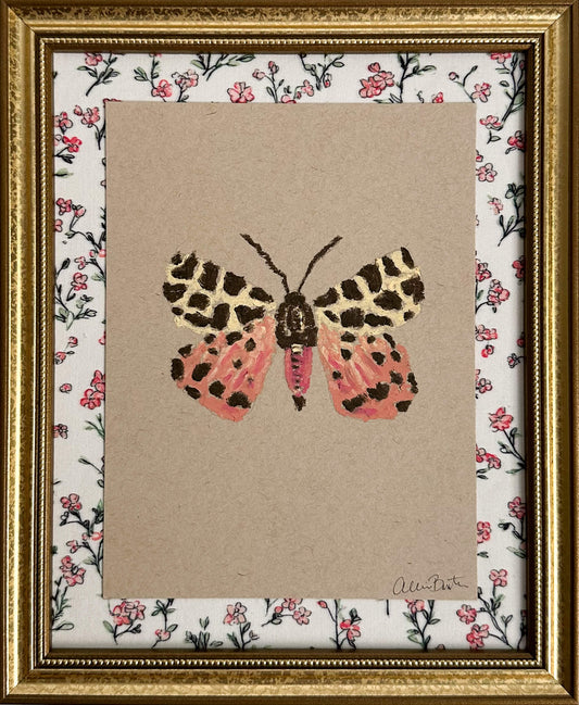 a pink, yellow and brown moth painted on tan paper. The paper is mounted on floral fabric and framed in a gold frame