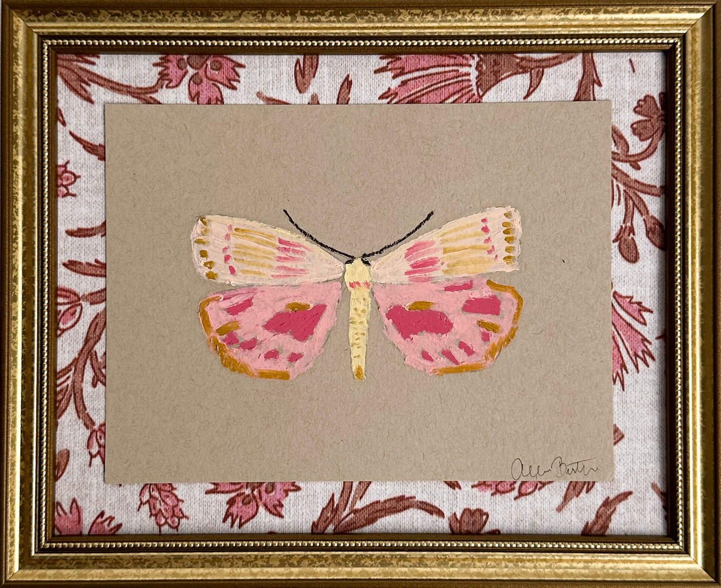 a pink and yellow butterfly painting on tan paper, mounted on pink and tan floral fabric and framed in a gold frame.