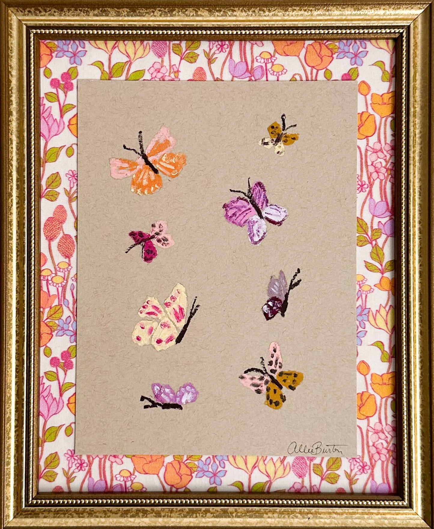 Eight colorful butterflies painted on tan toned paper and mounted on colorful, floral fabric. The painting is framed in a gold frame.