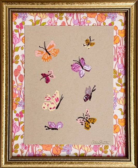 Eight colorful butterflies painted on tan toned paper and mounted on colorful, floral fabric. The painting is framed in a gold frame.