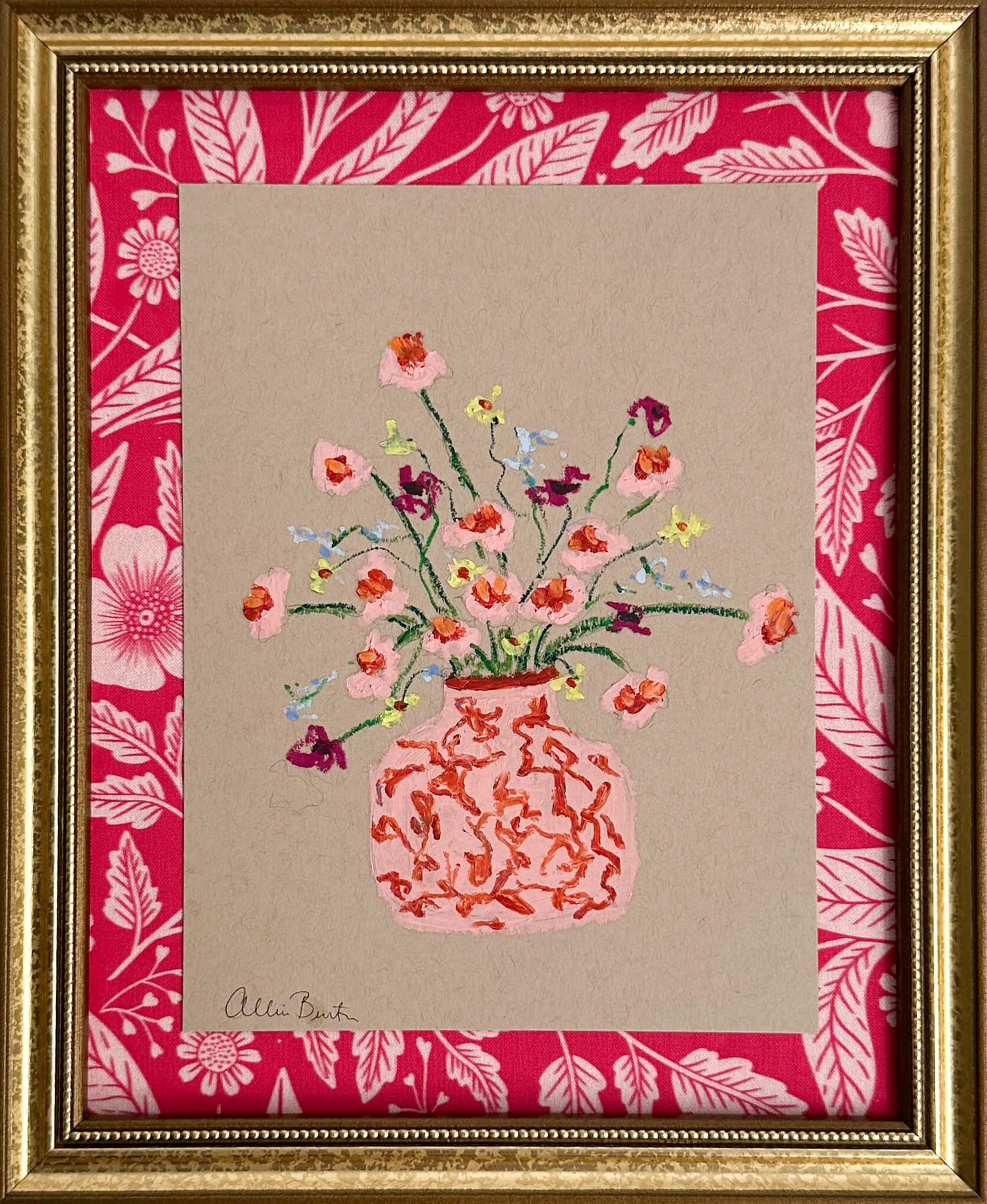A pink vase of small flowers painted on tan paper and mounted on pink floral fabric. The painting is framed in a gold frame.