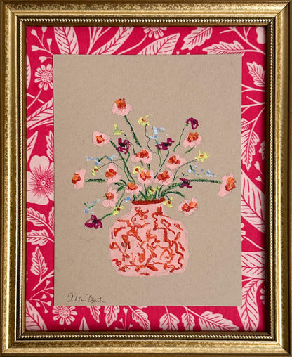 A pink vase of small flowers painted on tan paper and mounted on pink floral fabric. The painting is framed in a gold frame.