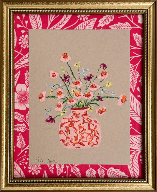 A pink vase of small flowers painted on tan paper and mounted on pink floral fabric. The painting is framed in a gold frame.