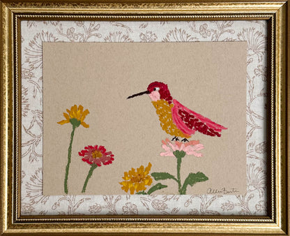 A painting of a colorful hummingbird and flowers on tan paper mounted on tan floral fabric. The painting is in a gold frame.