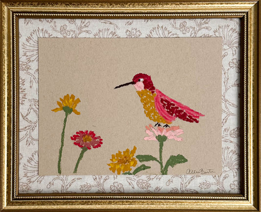 A painting of a colorful hummingbird and flowers on tan paper mounted on tan floral fabric. The painting is in a gold frame.