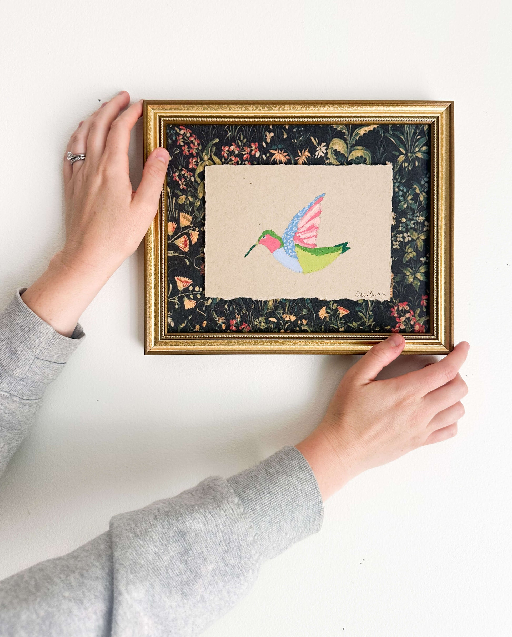 A painting on a colorful hummingbird on tan paper and mounted on dark floral paper. The painting is framed in a gold frame and shown on a white wall.