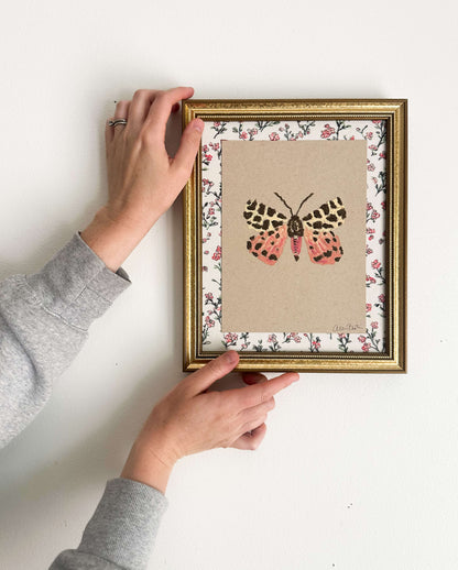 a pink, yellow and brown moth painted on tan paper. The paper is mounted on floral fabric, framed in a gold frame, and is shown on a white wall.