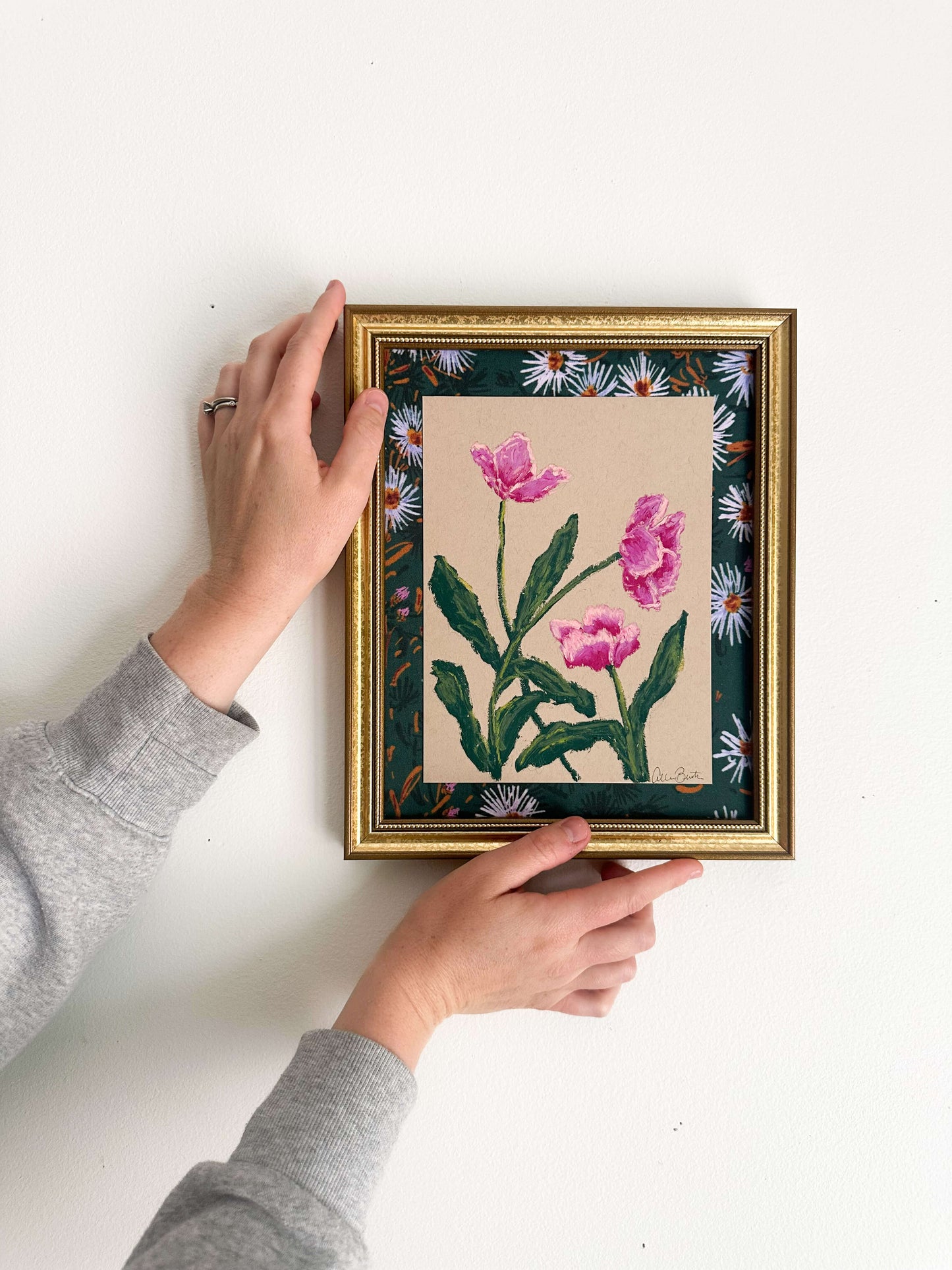 pink flowers painted on tan paper and mounted on floral fabric in a gold frame. The painting is shown on a white wall.
