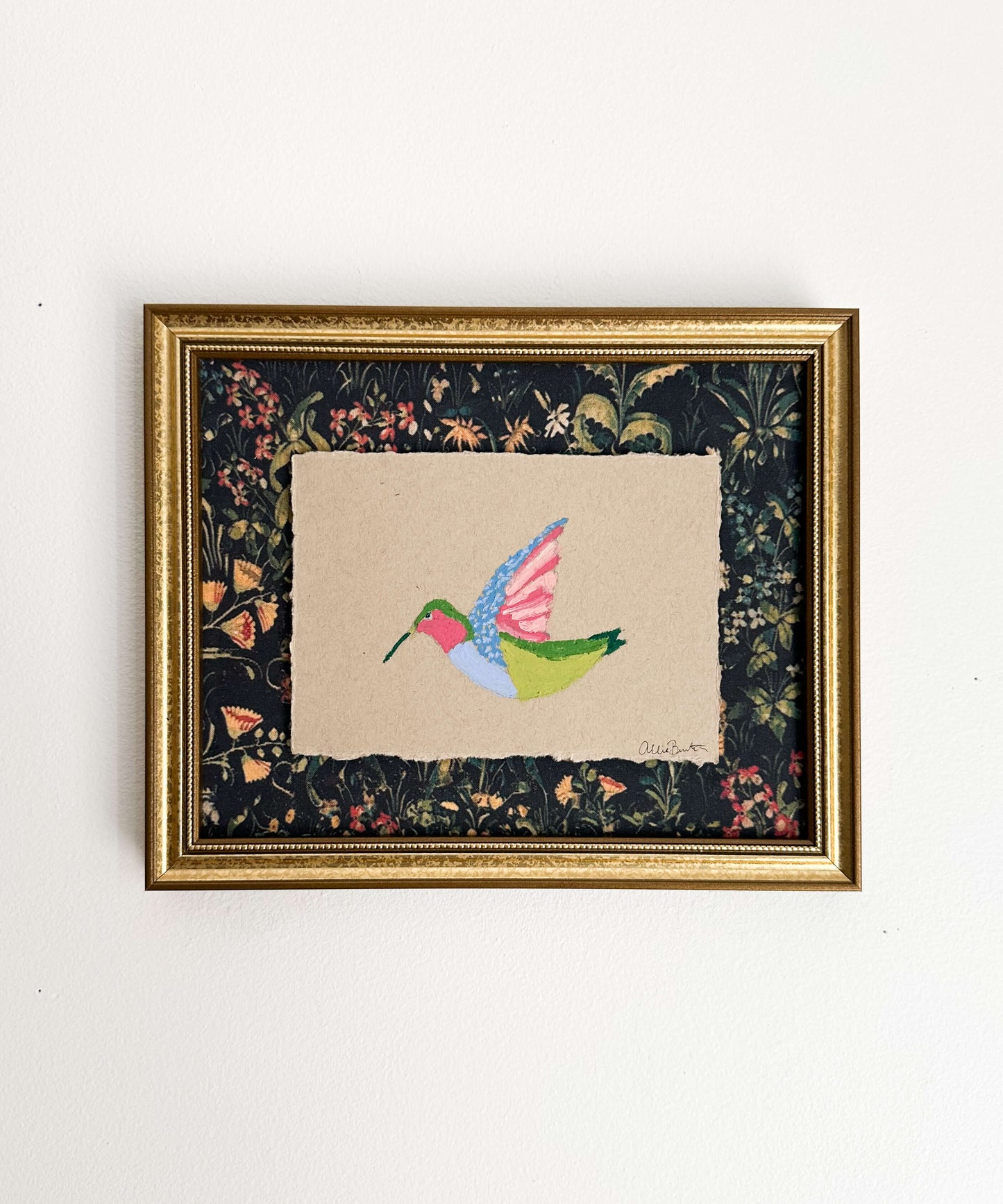 A painting on a colorful hummingbird on tan paper and mounted on dark floral paper. The painting is framed in a gold frame and shown on a white wall.