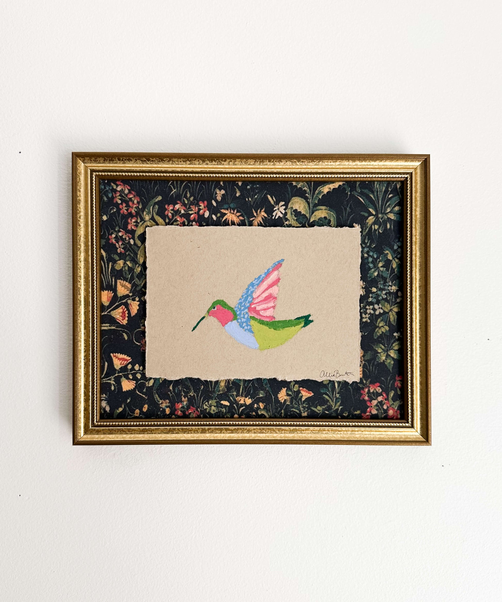 A painting on a colorful hummingbird on tan paper and mounted on dark floral paper. The painting is framed in a gold frame and shown on a white wall.