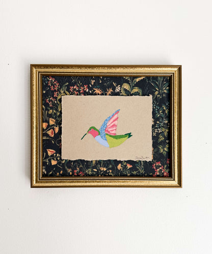 A painting on a colorful hummingbird on tan paper and mounted on dark floral paper. The painting is framed in a gold frame and shown on a white wall.