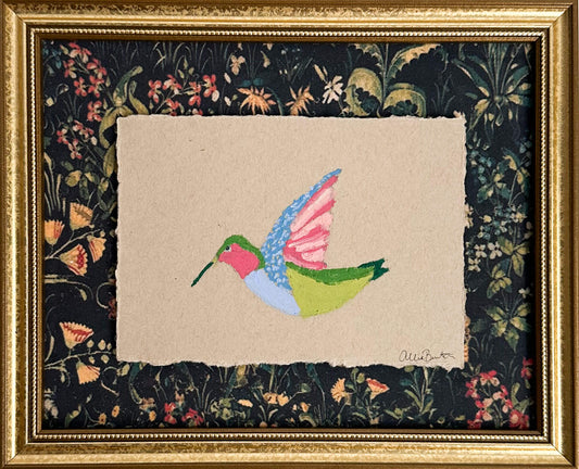 A painting on a colorful hummingbird on tan paper and mounted on dark floral paper. The painting is framed in a gold frame. 