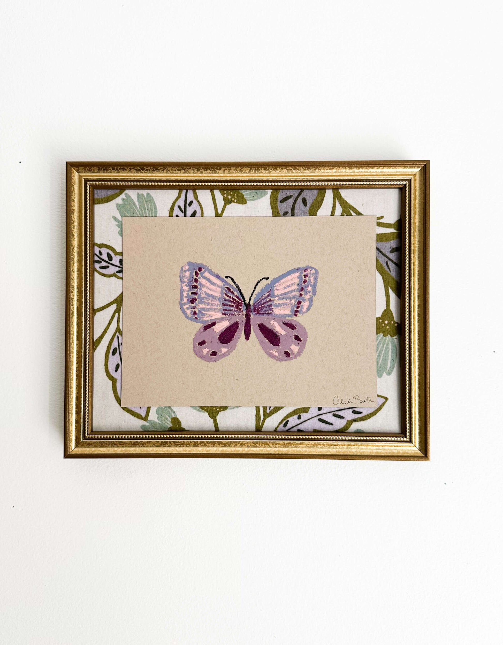 A painting of a purple butterfly on tan paper and mounted on floral fabric. The painting is in a gold frame and shown on a white wall.